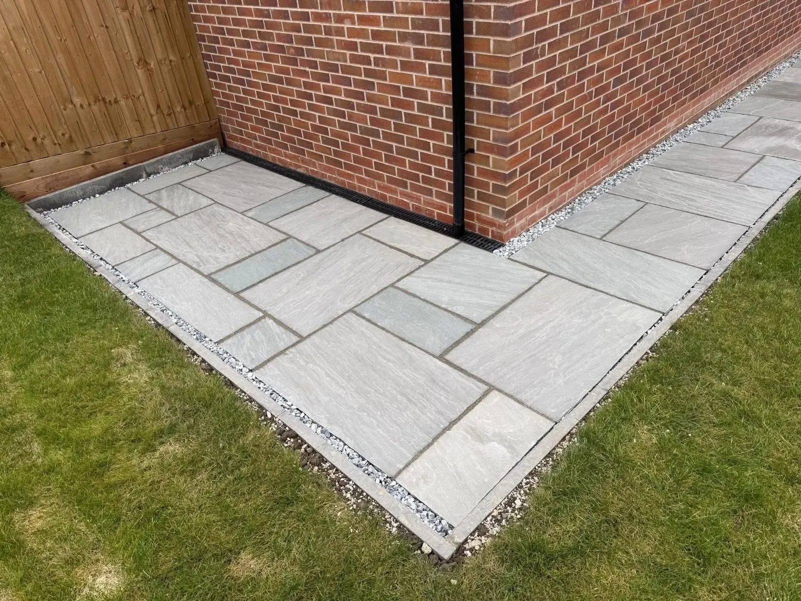 High-quality grey Indian sandstone paving around a house corner with a drainage channel and decorative gravel border.