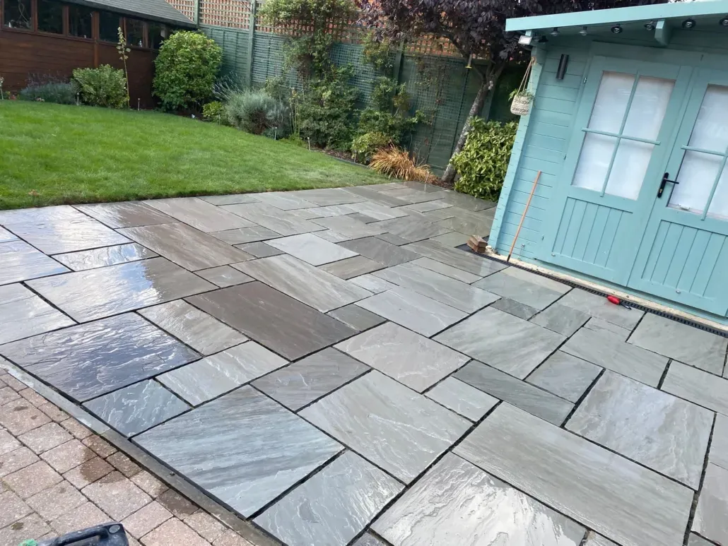 Recently completed Indian sandstone patio with grey and brown tones and varying slab sizes, next to a lawn and garden shed in Canterbury.