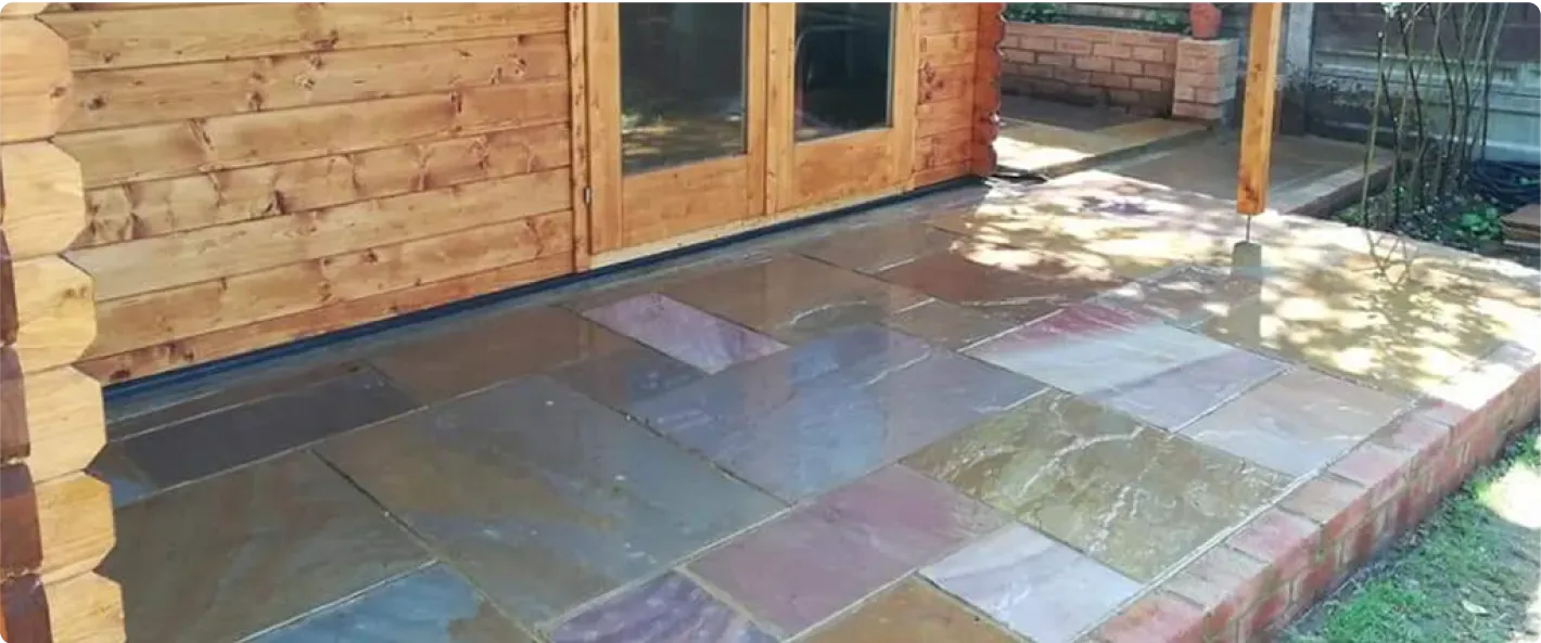 Newly laid multi-coloured Indian sandstone patio with brick edging next to a wooden shed or cabin, installed by Kent Paving & Resin LTD.