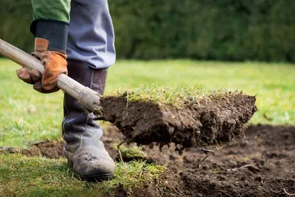 Close-up of someone wearing boots and gloves, using a shovel to remove turf and soil.